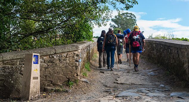 peregrinos caminando por un sendero del camino de santiago junto a un mojón con flecha amarilla